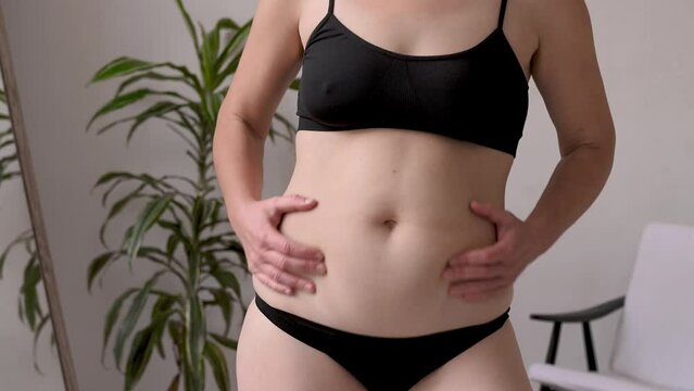 A woman in black underwear sorts out the folds on her stomach in front of a mirror after losing weight and dieting. Waist creases and cellulite. Excess weight