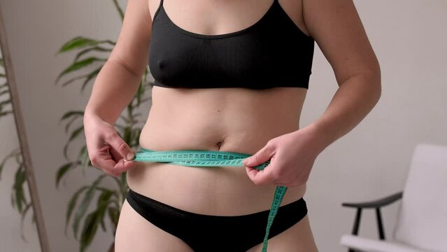 A woman in black lingerie sorts out the folds on her stomach in front of a mirror after losing weight and dieting. Measure your waist in centimetres. Waist creases and cellulite. Overweight and weight