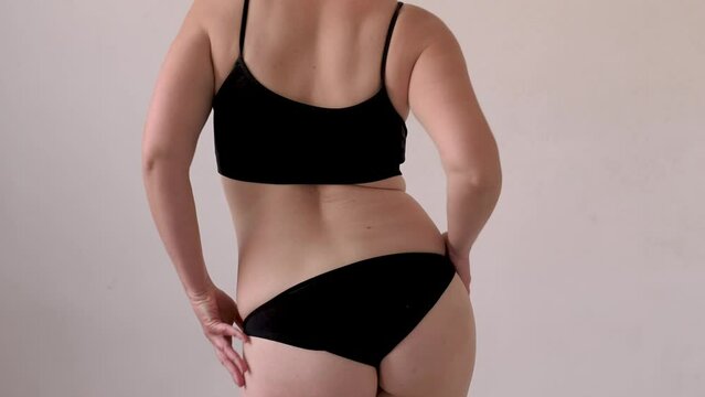 A woman in black lingerie sorts out the folds on her stomach after losing weight and dieting, dancing and twirling her hips. Waist creases and cellulite. Overweight and weight loss. View from the back