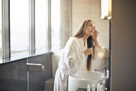 woman in a bathrobe in a hotel bathroom looks in the mirror and checks her skin