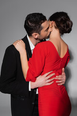 Elegant man kissing and hugging brunette girlfriend in red dress on grey background.