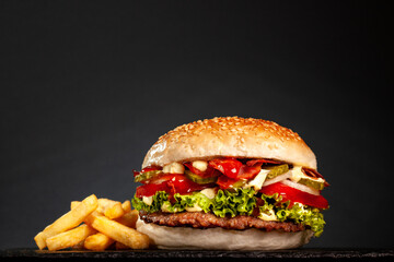 Bacon hamburger with onions tomatoes cucumber lettuce and french fries against dark gray background.