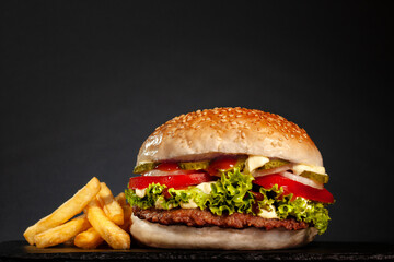Hamburger with onions tomatoes cucumber lettuce and french fries against dark gray background.