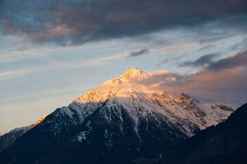 Ein verschneiter Berggipfel im goldenen Morgenlicht mit intensiven, grauen Wolken am Himmel