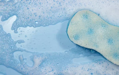 Tuinposter Blue sponge for bathing on soapy foam on the water on a blue background with copy space. Foam texture as background for image © Ruzanna