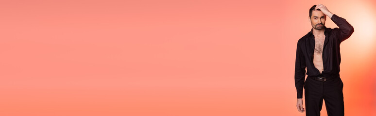 bearded man with hairy chest standing in black outfit and adjusting hair on pink, banner.