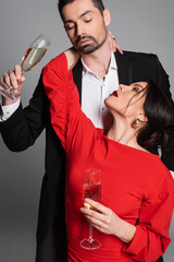 Seductive woman in red dress hugging boyfriend with champagne on grey background.