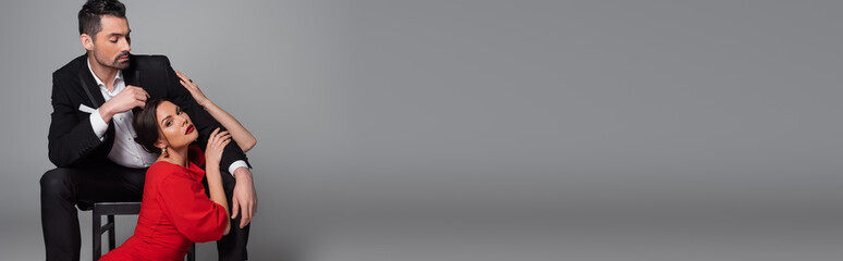 Sexy woman with burgundy lips touching boyfriend and looking at camera on grey background, banner.