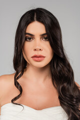 Naklejka premium brunette woman with shiny curly hair looking at camera isolated on grey.