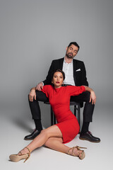 Seductive woman in dress and heels sitting near elegant boyfriend on chair on grey background.