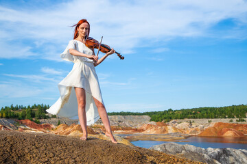 beautiful woman in elegant white dress and floral wreath playing violin on seashore