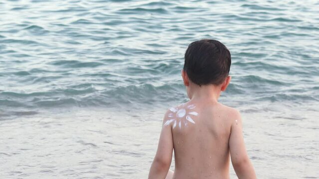 mother applying sun protection spf cream on child shoulder.kid boy wish sun drawn from white cream on back against sea on seashore coast line.happy child enjoy vacation sunblock lotion naked back view