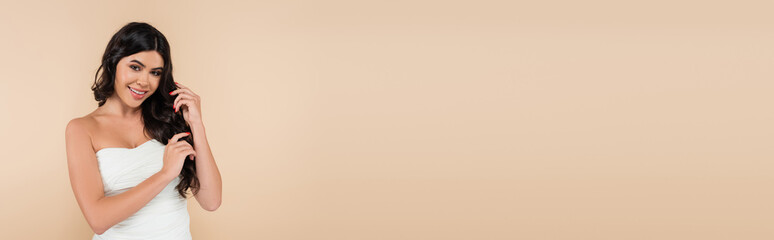 Fototapeta premium Positive brunette woman in top touching wavy hair isolated on beige, banner.