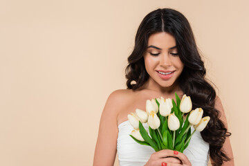 Positive brunette woman looking at white tulips isolated on beige.