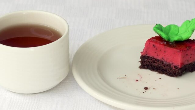 pastry piece slice of cake rectangle shape on white plate and black tea in cup.strawberry cake walnuts orange flower green leaf or pink macaron decoration.fork cutting the desert.tasty morning delight