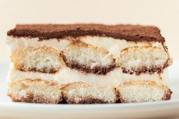 Homemade tiramisu cake on white plate against beige background. Traditional Italian no-bake dessert made of savoiardi, filled with mascarpone cheese, coffee espresso and sprinkled with cocoa powder