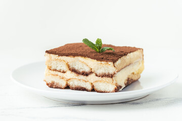 Homemade tiramisu cake on white plate against white background. Traditional Italian no-bake dessert made of savoiardi, filled with mascarpone cheese, coffee espresso and sprinkled with cocoa powder