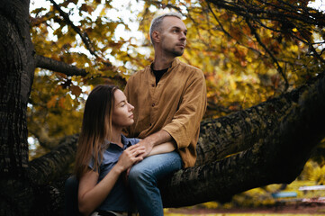 Young couple in a beautiful autumn park, with the man sitting on a tree and his girlfriend hugging his legs