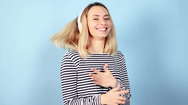 Cute energetic young blond woman with headphones dancing in good mood on isolated blue background Charming smiling female having fun alone at studio