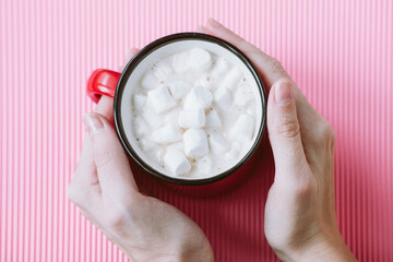 Fototapeta na wymiar The girl's hands hold a large cup of cocoa or hot chocolate with marshmallows on a pink background, top view