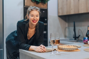 Beautiful dressed woman drinking champagne near the Christmas table. Stylish adult girl in shiny black clothes celebrates new year at home. People celebrate New Year in the living room.