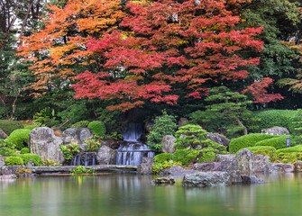 fukuoka, kyushu - december 07 2022: Beautiful autumn landscape with red momiji maple trees in rain overlooking the Sandan-Ochi-no-Taki Waterfall in the Ue-no-Ike pond of the Japanese Ohori garden.