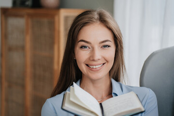Obraz premium Close up cheerful caucasian woman in blue shirt holds opened diary looks at camera toothy smiles at home. Adorable Italian girl offers book. Pretty blonde female planning week. Successful people.