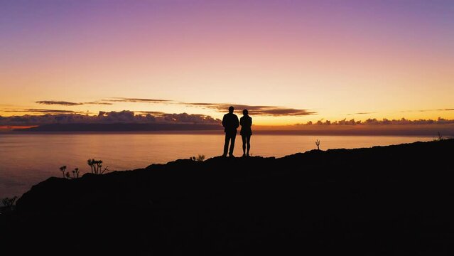 Couple silhouette man and woman two tourists stand on the top of mountain during colorful sunset twilight. Night nature. Aerial view of glimmering pacific beach. Friendly family successful teamwork.