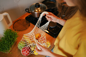 A young woman unpacks a mesh bag with fresh produce in her kitchen. The concept of ecological consumption