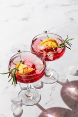 Two glass of pomegranate iced cocktail close up with rosemary and lemon on white marble background isolated with clear shadows in harsh light. Festive drink, valentines day concept. Text space