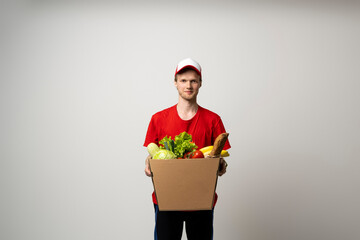 Delivery Concept. Smiling young delivery man in red uniform carrying package box of grocery food and drink from store, market on white studio background.