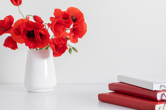 White office desk, stack of notebooks on table, vase with red flowers poppies. Front view. Place for text, copy space, mockup