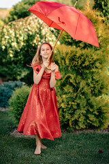 A little girl plays with an umbrella in a red dress on a green field near the house