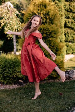 A little girl in a red long dress is dancing on a green plot of land near her country house