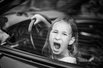 A little girl is sitting behind the wheel of a car, messing around and yelling at the photographer....