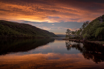 Sunset with orange clouds reflecting on water surface of lake in Southern Norway in summer