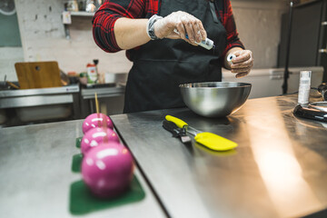Baker wearing transparent gloves, black apron and red plaid shirt putting blue food coloring into the metal bowl. Yellow spatula placed next to the metal bowl. Three cakes decorated with pink icing on