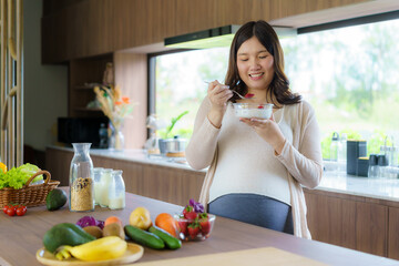 Beautiful Asian pregnant woman eating with lust cereals on breakfast in kitchen at home, enjoying...