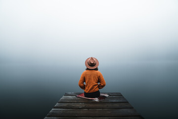 Back view of fashioned young woman sitting on wooden dock looking at view on a misty morning....