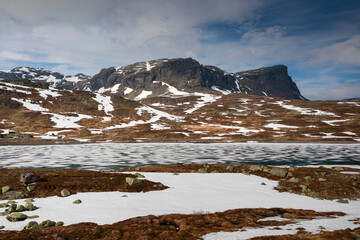 Snowy landscape of Hardangervidda with mountains and icy lake in Norway, red hut at the lakeside