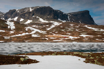 Snowy landscape of Hardangervidda with mountains and icy lake in Norway, red hut at the lakeside