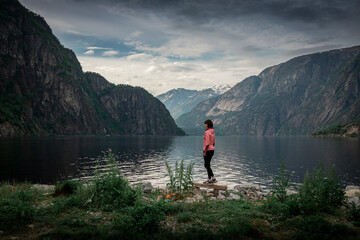 Woman standing at waterfront of lake in the mountain landscape Eidfjord in Norway, looking into the fjord, clouds in the sky