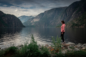 Fototapeta na wymiar Woman standing at waterfront of lake in the mountain landscape Eidfjord in Norway, looking into the fjord, clouds in the sky