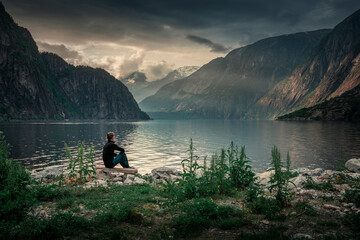 Man sitting at waterfront of lake in the mountain landscape Eidfjord in Norway, looking into the fjord, clouds in the sky during sunset