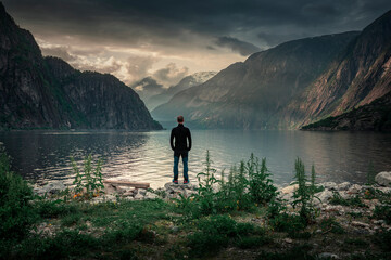 Fototapeta na wymiar Man standing at waterfront of lake in the mountain landscape Eidfjord in Norway, looking into the fjord, clouds in the sky during sunset