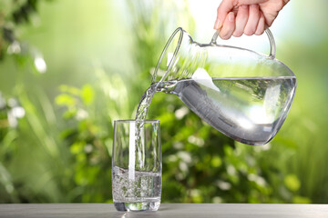 Woman pouring water from jug into glass on light grey table outdoors, closeup
