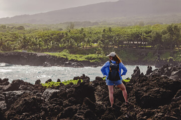 Woman with a backpack standing on the black rock lava beach, looking on the ocean