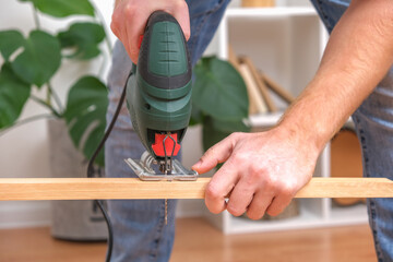 The hands of a master carpenter with an electric jigsaw in his hands cutting off a piece of wood. Male hands using fret saw for cutting wood. A carpenter cuts wood with jigsaw in a home workshop.