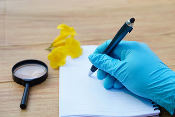 Closeup  hands in protective gloves hold magnifying glass to inspect flowers, holds pen to write on paper. Concept, science subject, project work. Experiment, education, learning by doing approach.   