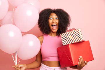 Positive overjoyed dark curly haired young woman, holds gifts and balloons, poses over pink wall